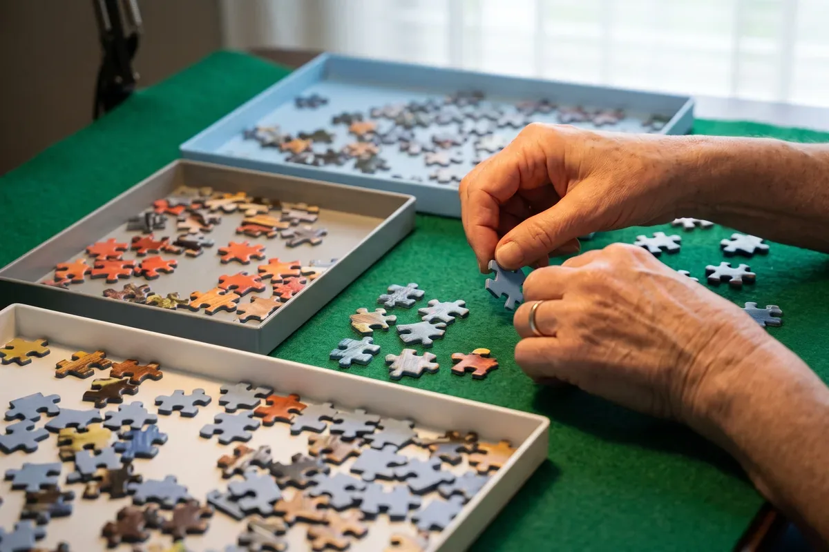 Older woman's hands sorting large puzzle pieces into shallow trays on a felt mat under a desk lamp